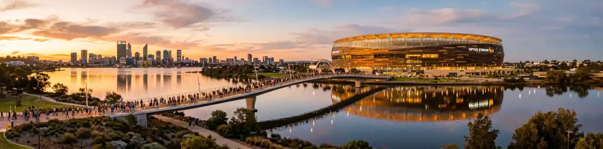 Optus Stadium Perth at sunset viewed from across the Swan River with the Matagarup Bridge and bronze facade glowing in golden hour light